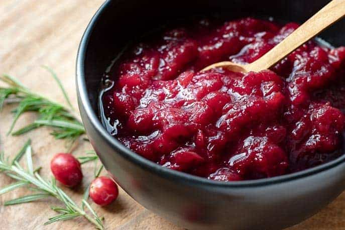 Cranberry sauce with wooden spoon in a black bowl with rosemary and cranberries on a board.