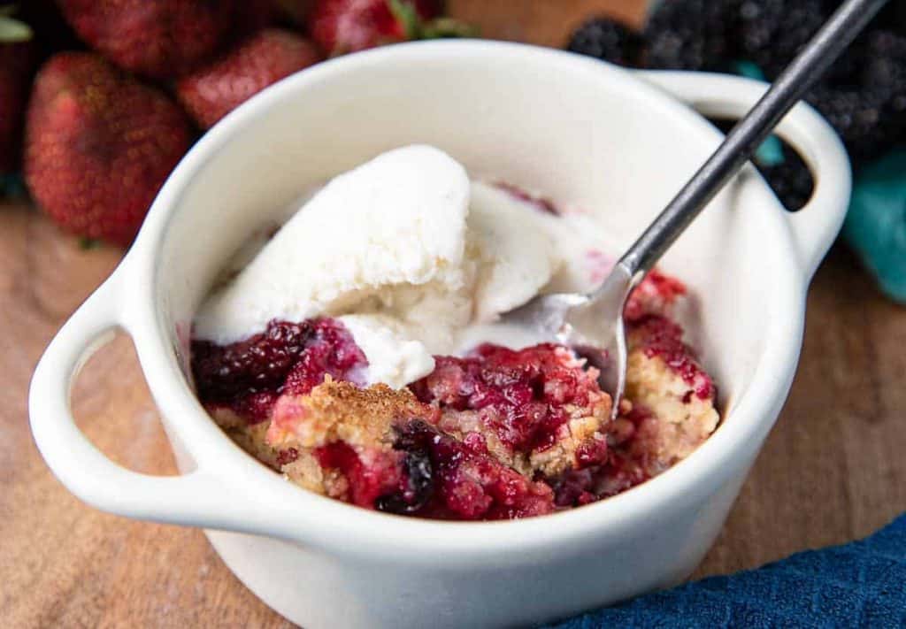 small white dish with handles with mixed berry cobbler and a black spoon, strawberries and blackberries in background