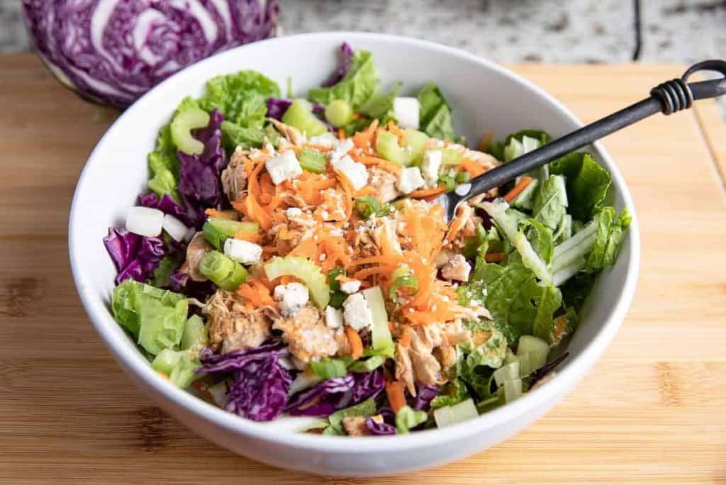 white bowl with black spoon on a wooden board with buffalo chicken, cabbage, celery, blue cheese, green onions, cabbage and carrots